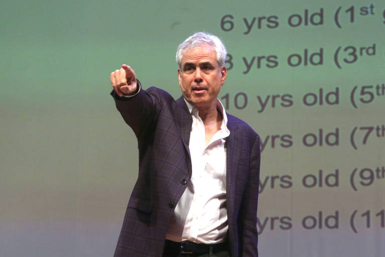 Um homem de cabelo grisalho e com uma expressão séria está em pé em um palco, apontando com a mão direita. Ele usa um paletó escuro e uma camisa branca. Ao fundo, há uma projeção com texto que menciona idades de 6 a 11 anos, indicando diferentes graus ou níveis.