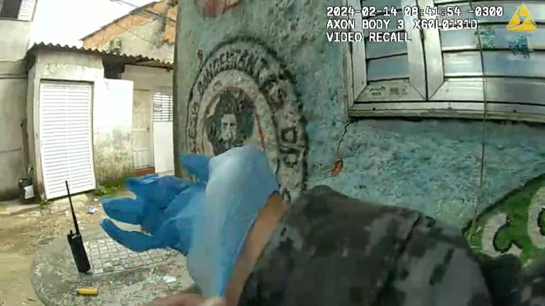 The image shows a hand putting on a blue glove, with a background that includes a green wall with a graffiti design and a window with silver blinds. The setting is an outdoor area, possibly a patio or backyard, with a visible wall structure and a black two-way radio on top of a round concrete table.