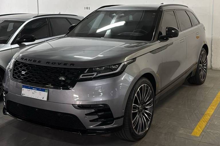 A imagem mostra um SUV de cor prata estacionado em um estacionamento. O veículo possui rodas grandes e um design moderno. Ao fundo, há outros carros, e o ambiente é bem iluminado, com paredes brancas e piso cinza.
