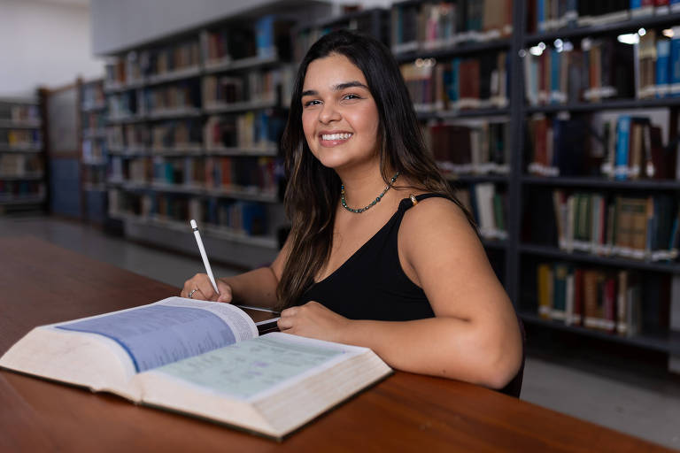 Uma jovem mulher está sentada em uma mesa de biblioteca, sorrindo enquanto escreve em um caderno. Ela tem cabelos longos e escuros, e usa uma blusa preta. Na mesa, há um grande livro aberto, com páginas azuis e verdes. Ao fundo, prateleiras cheias de livros estão visíveis, indicando que o ambiente é uma biblioteca.