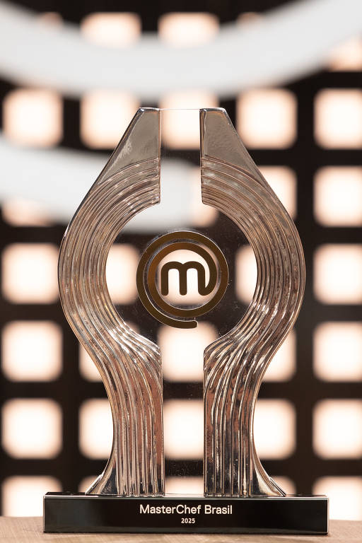 A imagem mostra um troféu do MasterChef Brasil, que é feito de um material transparente e possui um design moderno. O troféu tem uma base retangular e uma forma que se assemelha a duas mãos unidas, com um círculo no centro que contém a letra 'm'. Abaixo do círculo, está escrito 'MasterChef Brasil 2023'. O fundo da imagem é desfocado, com luzes que criam um efeito visual interessante.