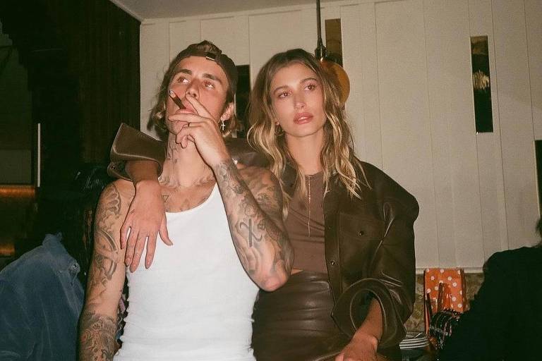 A imagem mostra um homem e uma mulher em um ambiente social. O homem está usando uma camiseta branca e calças escuras, com tatuagens visíveis nos braços. Ele está segurando um cigarro e tem uma expressão relaxada. A mulher está ao seu lado, vestindo um vestido curto e elegante, com cabelo solto. O fundo apresenta uma decoração de bar ou restaurante, com luzes suaves e móveis de madeira.