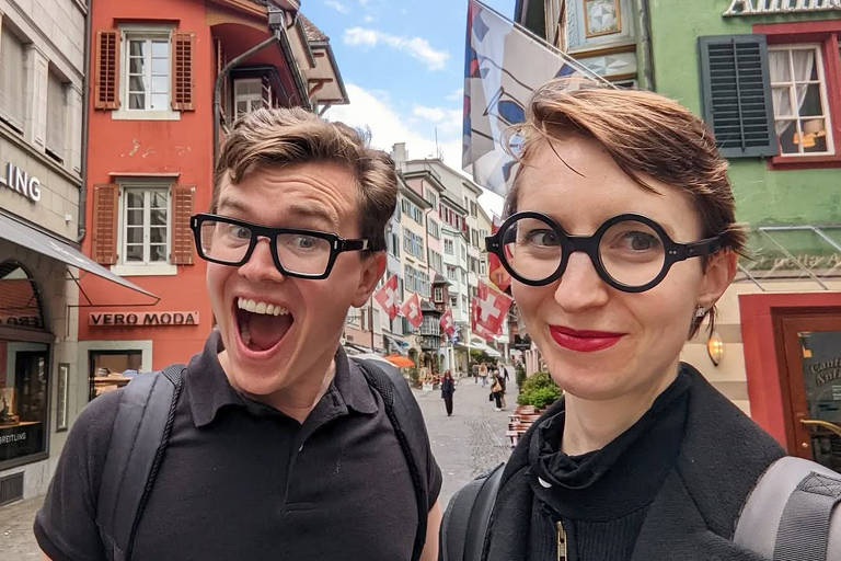 Um casal está posando para a foto em uma rua charmosa e colorida. O homem à esquerda está sorrindo amplamente, usando uma camiseta preta e óculos grandes. A mulher à direita tem um olhar sorridente, com cabelo curto e usa óculos redondos. Ao fundo, há edifícios com fachadas coloridas e bandeiras penduradas, sugerindo um ambiente turístico.
