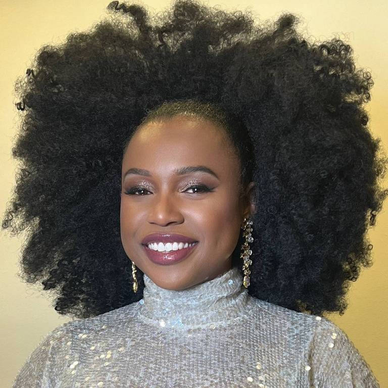 A imagem mostra uma mulher sorridente com cabelo afro volumoso e bem definido. Ela está usando uma blusa prateada com brilho e brincos longos. O fundo é de uma cor clara, destacando sua aparência.