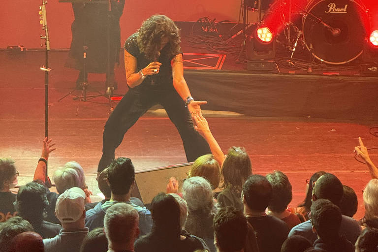 A imagem mostra um cantor se apresentando em um palco, com uma iluminação suave em tons de vermelho e laranja. O artista está agachado, interagindo com a plateia, que está bem próxima e visivelmente animada. Ao fundo, há uma bateria e outros músicos, enquanto o público, composto por várias pessoas, observa atentamente. Algumas pessoas estão segurando celulares para registrar o momento.