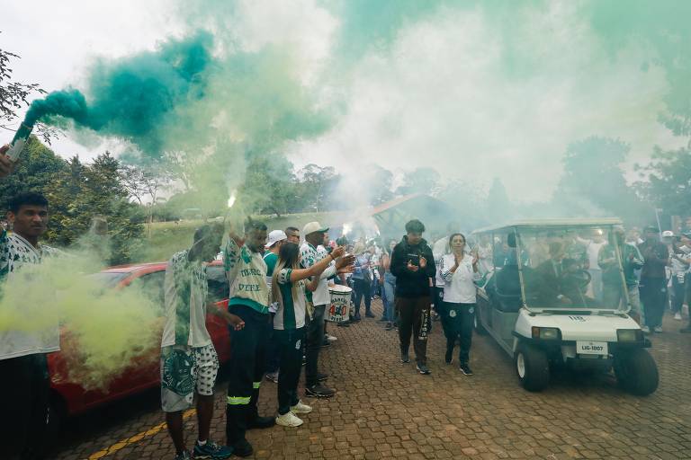 Um grupo de pessoas está reunido em um enterro, segurando sinalizadores que emitem fumaça verde. Eles estão em uma área externa, com árvores ao fundo. Algumas pessoas estão vestindo camisetas e bonés, enquanto outras estão em um veículo elétrico
