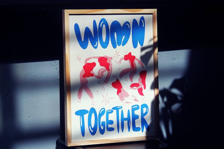 A imagem mostra um pôster emoldurado com a frase "WOMEN TOGETHER". As palavras estão escritas em letras grandes, com "WOMEN" em azul e "TOGETHER" em azul também. O fundo do pôster apresenta ilustrações de mulheres em várias poses, em tons de vermelho e rosa. O pôster está apoiado em uma superfície escura, e há sombras projetadas ao redor, criando um contraste com o ambiente iluminado.