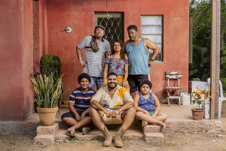 A imagem mostra uma família composta por seis pessoas, posando em frente a uma casa de paredes vermelhas. Na parte inferior, um homem está sentado, enquanto cinco pessoas estão em pé atrás dele. As crianças estão vestidas com camisetas coloridas, e os adultos usam roupas casuais. Ao lado da casa, há plantas em vasos e uma cadeira branca visível ao fundo.