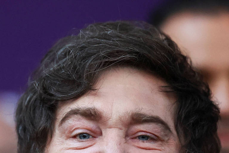 La imagen muestra a un hombre sonriente con cabello castaño y ondulado. Tiene ojos claros y una expresión amigable. El fondo es de color morado y hay otras personas borrosas en el fondo.