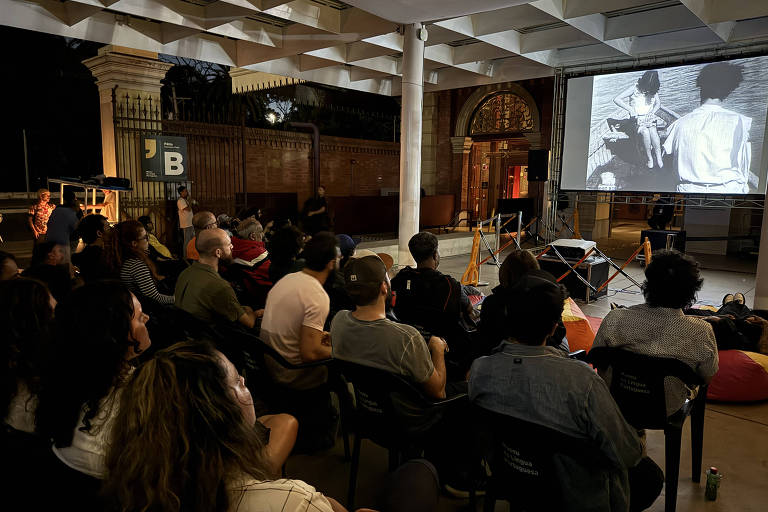 A imagem mostra um grupo de pessoas assistindo a uma exibição de filme ao ar livre. O ambiente é noturno, com uma tela grande projetando uma cena em preto e branco. As pessoas estão sentadas em cadeiras, algumas em almofadas, e parecem atentas ao que está sendo exibido. O local tem uma arquitetura com colunas e um ambiente acolhedor.
