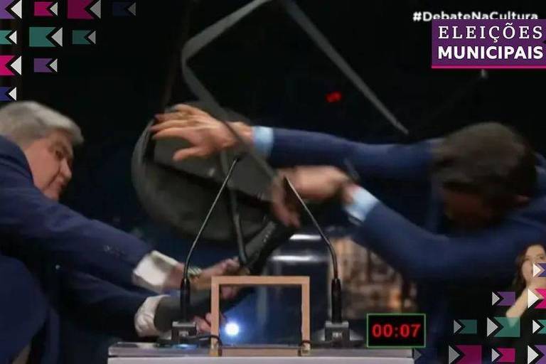 A imagem mostra dois homens em um debate, aparentemente em um momento de conflito. Um dos homens está tentando puxar um objeto, enquanto o outro tenta segurá-lo. O ambiente parece ser um estúdio de televisão, com um cronômetro visível na parte inferior da imagem, marcando 00:07. Há elementos gráficos ao redor, indicando que se trata de um debate sobre eleições municipais.