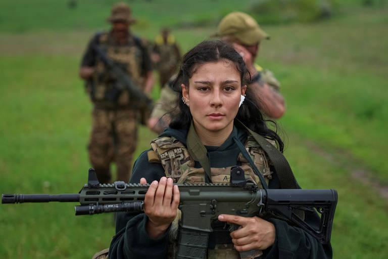 A imagem mostra uma jovem mulher vestindo um uniforme militar e segurando um fuzil. Ela tem cabelos longos e escuros, e está em um campo aberto com grama verde ao fundo. Ao fundo, há outros dois soldados, um deles com um chapéu e outro com um fuzil, que parecem estar se preparando para uma missão. A expressão da mulher é séria e focada.