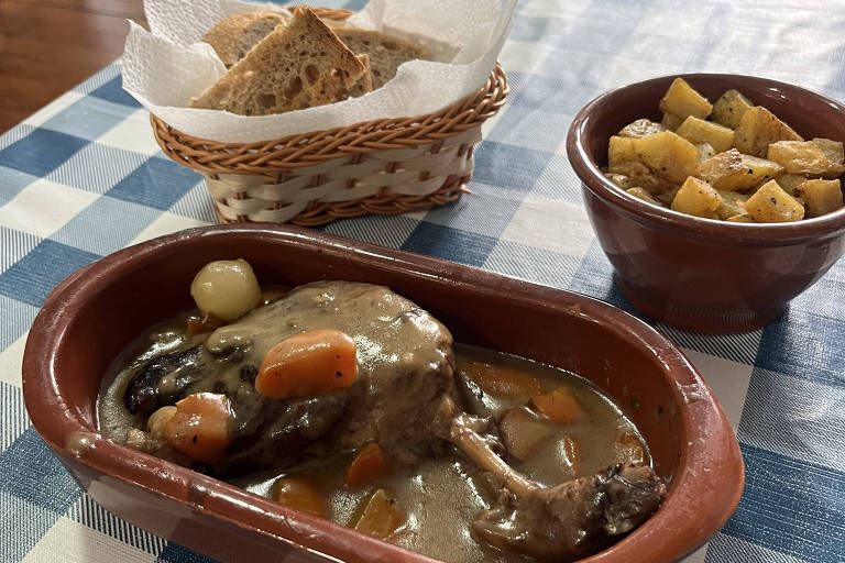 A imagem mostra um prato de comida caseira em uma mesa com toalha xadrez azul e branca. O prato principal é uma carne com molho, acompanhada de cenouras e cebolas, servida em uma travessa de barro. Ao lado, há uma tigela com batatas fritas e uma cesta com pão. O ambiente parece ser um restaurante aconchegante.