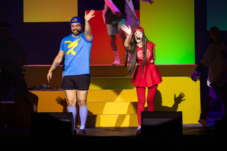 Uma apresentação no palco com duas pessoas. À esquerda, um homem vestido com uma camiseta azul e shorts, acenando. À direita, uma mulher vestindo um vestido vermelho e meias vermelhas, também acenando. O fundo do palco é colorido, com luzes em tons de verde, amarelo e roxo. Há outras pessoas ao fundo, mas não estão claramente visíveis.