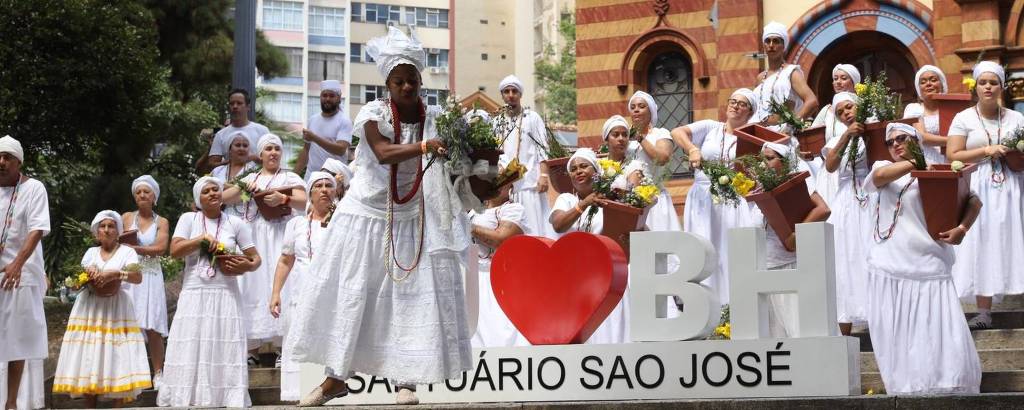 A imagem mostra um grupo de pessoas vestidas com roupas brancas, algumas com turbantes, realizando uma celebração em um espaço público. Elas estão em frente a um letreiro que diz 'I ❤️ BH' e 'Praça São José'. Algumas pessoas seguram vasos com flores, enquanto outras estão em poses de dança ou celebração. Ao fundo, há um edifício com arquitetura colorida e moderna, e árvores ao redor.