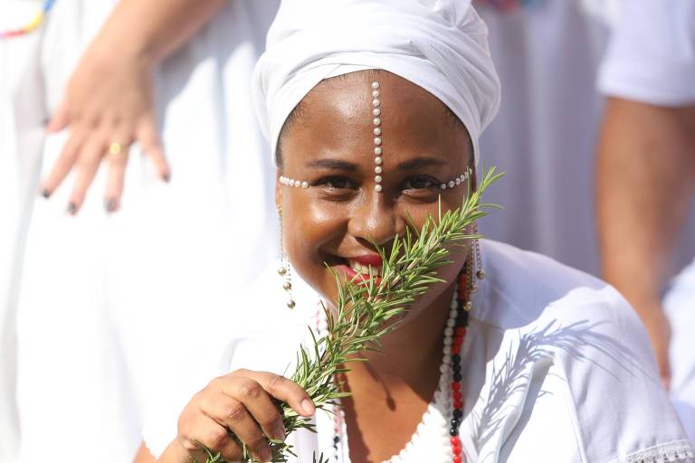 Uma mulher sorridente, vestindo roupas brancas e um turbante branco, segura um ramo de erva em sua mão. Ela tem adornos de pérolas no rosto e um colar colorido. Ao fundo, outras pessoas também vestem roupas brancas, sugerindo um evento cultural ou religioso.