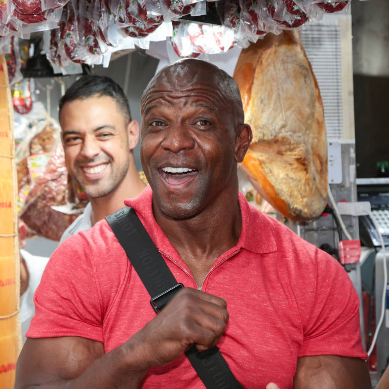 A imagem mostra dois homens sorrindo em um mercado. O homem à frente, com pele escura, está usando uma camiseta vermelha e tem um sorriso largo. Ele parece estar se divertindo. O homem ao fundo, com pele clara, também está sorrindo e parece estar em um ambiente de mercado, com produtos pendurados ao fundo, incluindo carnes. A atmosfera é alegre e descontraída.