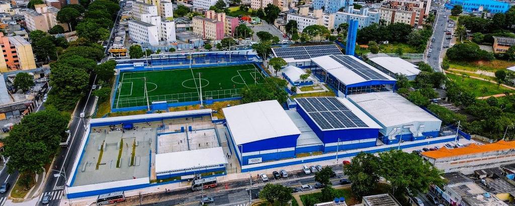 A imagem mostra uma vista aérea de um complexo esportivo. O local possui um campo de futebol verde, cercado por uma grade, e várias edificações ao redor, com telhados azuis e brancos. Há também áreas de quadras esportivas e um estacionamento visível na parte inferior da imagem. Ao fundo, pode-se ver uma área urbana com prédios e árvores.