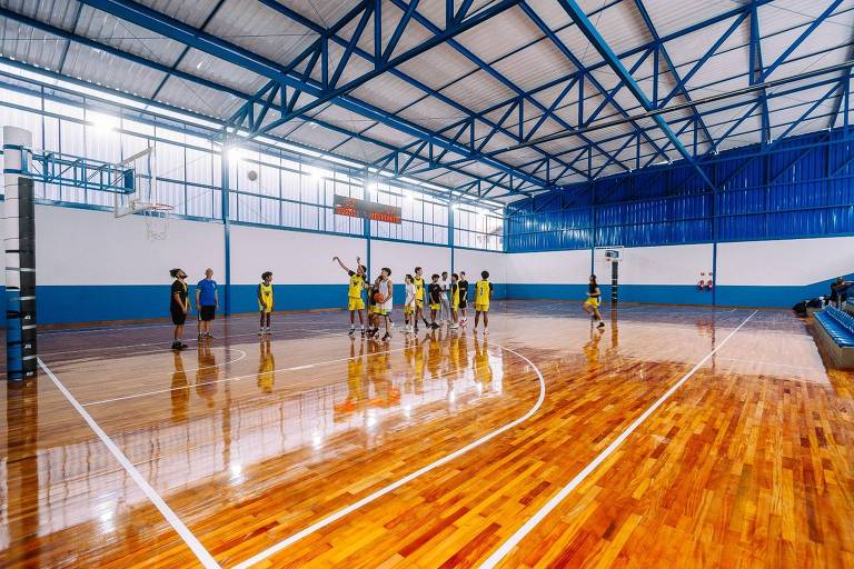 A imagem mostra uma quadra de basquete coberta com piso de madeira brilhante. Há várias pessoas jogando basquete, algumas vestindo uniformes amarelos e outras com uniformes escuros. O ambiente é bem iluminado, com janelas grandes ao fundo e uma estrutura metálica azul no teto. Há também uma tabela de basquete visível em uma das extremidades da quadra.