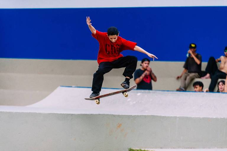 Um jovem skatista está realizando um truque no ar sobre uma rampa em um parque de skate. Ele veste uma camiseta vermelha e calças escuras, enquanto se prepara para aterrissar. Ao fundo, há um grupo de pessoas assistindo ao desempenho, algumas delas aplaudindo. O cenário é composto por uma parede azul e uma superfície de skate clara.