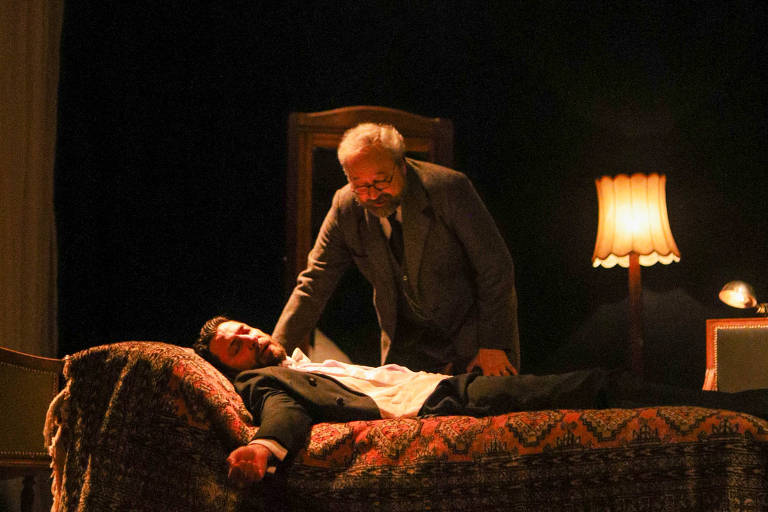 A imagem mostra uma cena de teatro com dois personagens. Um homem mais velho, vestido com um terno, está inclinado sobre um homem mais jovem que está deitado em uma cama. O jovem parece estar inconsciente ou em um estado de fraqueza. O ambiente é iluminado por uma lâmpada de pé e um lustre acima, criando uma atmosfera dramática. Ao fundo, há um espelho e cortinas que sugerem um cenário de quarto.