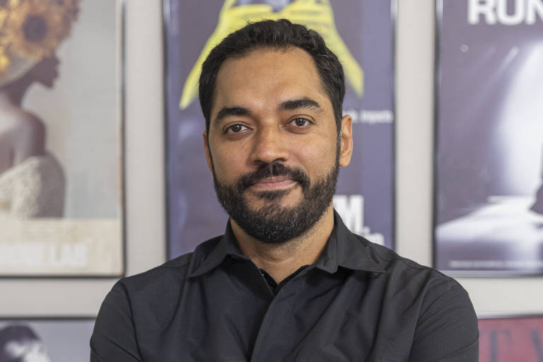 Um homem de pele morena, com barba e cabelo curto, está posando em pé com os braços cruzados. Ele usa uma camisa preta e um relógio no pulso. Ao fundo, há uma parede decorada com vários pôsteres de filmes, apresentando imagens em preto e branco e cores vibrantes. O ambiente parece ser um espaço cultural ou artístico.