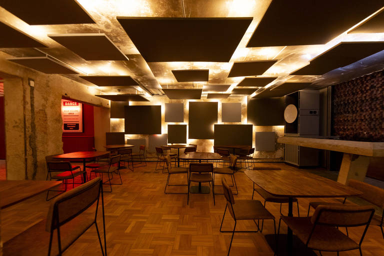 A imagem mostra um ambiente interno de um café moderno. O espaço possui mesas e cadeiras de metal com tampo de madeira, dispostas de forma organizada. O teto é decorado com painéis acústicos pretos e há iluminação suave que destaca o ambiente. As paredes têm um acabamento em concreto e há uma área com um fundo vermelho. No fundo, é possível ver um balcão e um eletrodoméstico.