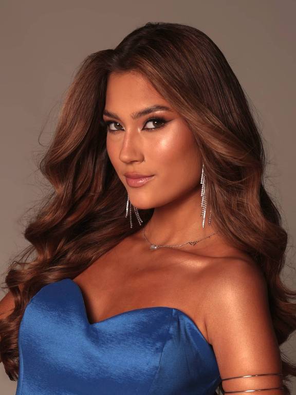A imagem mostra uma mulher com cabelo longo e ondulado, usando um vestido azul. Ela está posando de lado, com um olhar confiante. A maquiagem é bem elaborada, destacando os olhos e os lábios. A mulher também usa brincos longos e brilhantes. O fundo é neutro, destacando a figura dela.