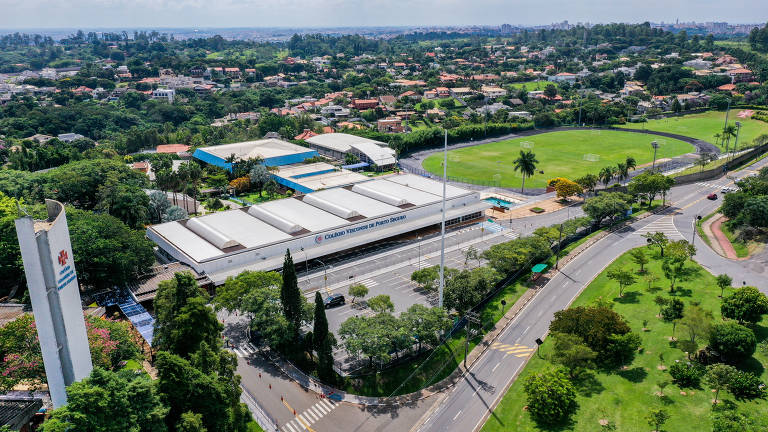 A imagem mostra uma vista aérea de um complexo esportivo, com um campo de grama verde ao fundo e várias estruturas de edifícios ao redor. Há árvores e áreas verdes nas proximidades, além de uma estrada que contorna o local. O céu está claro e a cidade pode ser vista ao fundo, com várias construções e vegetação.