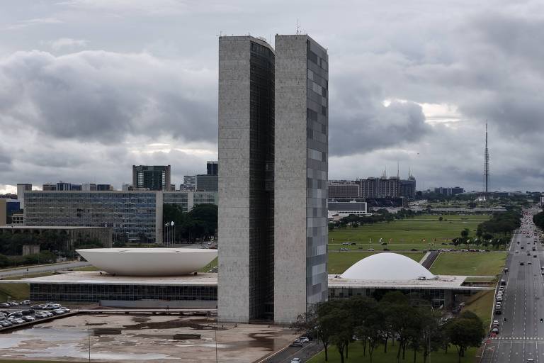 A imagem mostra o Congresso Nacional do Brasil, localizado em Brasília. O edifício é composto por duas torres retangulares e duas cúpulas, uma cúpula convexa e outra côncava. O céu está nublado, e há uma estrada em primeiro plano com alguns carros. Ao fundo, é possível ver outros edifícios e uma antena.