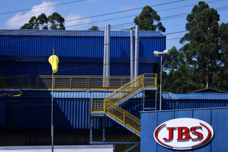 A imagem mostra uma instalação da JBS, com um grande edifício azul ao fundo. Há uma escada amarela visível na lateral do edifício e um balão amarelo preso em um mastro. O logotipo da JBS está em destaque na parte inferior direita da imagem.