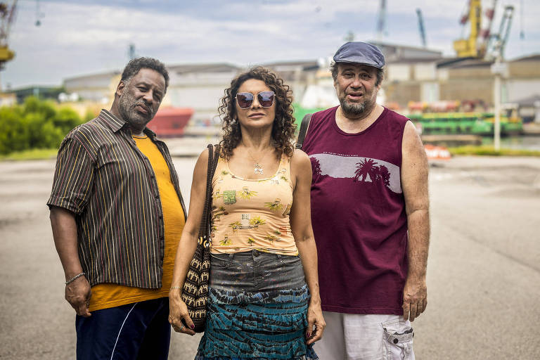 Três pessoas estão em pé em um ambiente urbano, possivelmente próximo a um porto. À esquerda, um homem de pele escura, com cabelo curto e barba, usa uma camisa listrada e calças escuras. No centro, uma mulher de pele clara, com cabelo cacheado e óculos escuros, veste uma blusa amarela com estampas florais e uma saia longa. À direita, um homem de pele clara, com cabelo curto e barba, usa uma camiseta roxa e calças brancas. Ao fundo, há estruturas industriais e equipamentos de porto.