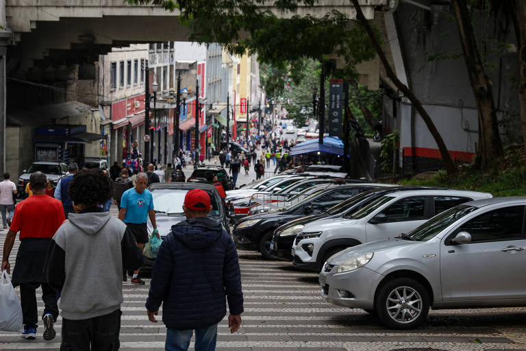 A imagem mostra uma rua movimentada em um ambiente urbano. No primeiro plano, há várias pessoas caminhando, algumas carregando sacolas. Ao lado, há uma fila de carros estacionados. Ao fundo, é possível ver mais pessoas e lojas, com uma atmosfera de cidade. O céu está nublado, sugerindo um dia sem sol
