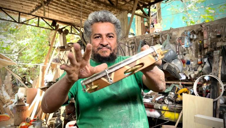 Um homem com cabelo grisalho e barba, vestindo uma camiseta verde, está em uma oficina de marcenaria. Ele segura uma ferramenta de madeira com uma mão e faz um gesto com a outra, como se estivesse mostrando a ferramenta. Ao fundo, há várias ferramentas e materiais de marcenaria, além de luz natural entrando pela janela.