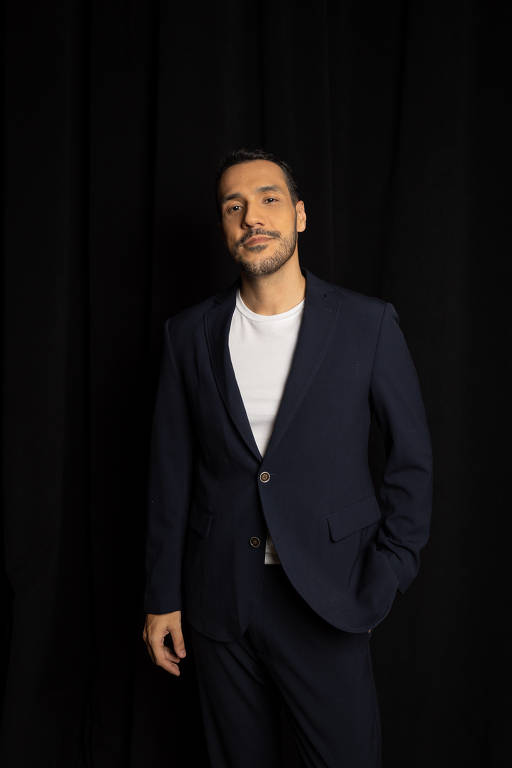 Um homem está posando em um fundo preto. Ele usa um terno azul escuro com uma camisa branca por baixo. O homem tem uma expressão confiante e está com uma mão no bolso do terno, enquanto a outra está relaxada ao lado do corpo. Seu cabelo é curto e bem arrumado.