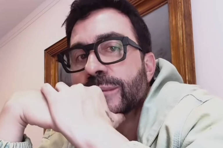 Um homem com barba e óculos está posando para a câmera. Ele usa uma jaqueta clara e está com os braços cruzados, apoiando o queixo na mão. Ao fundo, há uma parede clara e um espelho emoldurado em madeira.