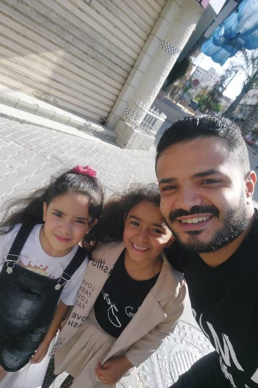 A imagem mostra um homem sorridente posando para a câmera ao lado de duas meninas. As meninas estão vestidas de forma casual, uma delas usa um vestido claro e a outra uma camiseta com suspensórios. O fundo apresenta uma rua com calçamento e edifícios ao fundo, além de árvores e um céu claro.