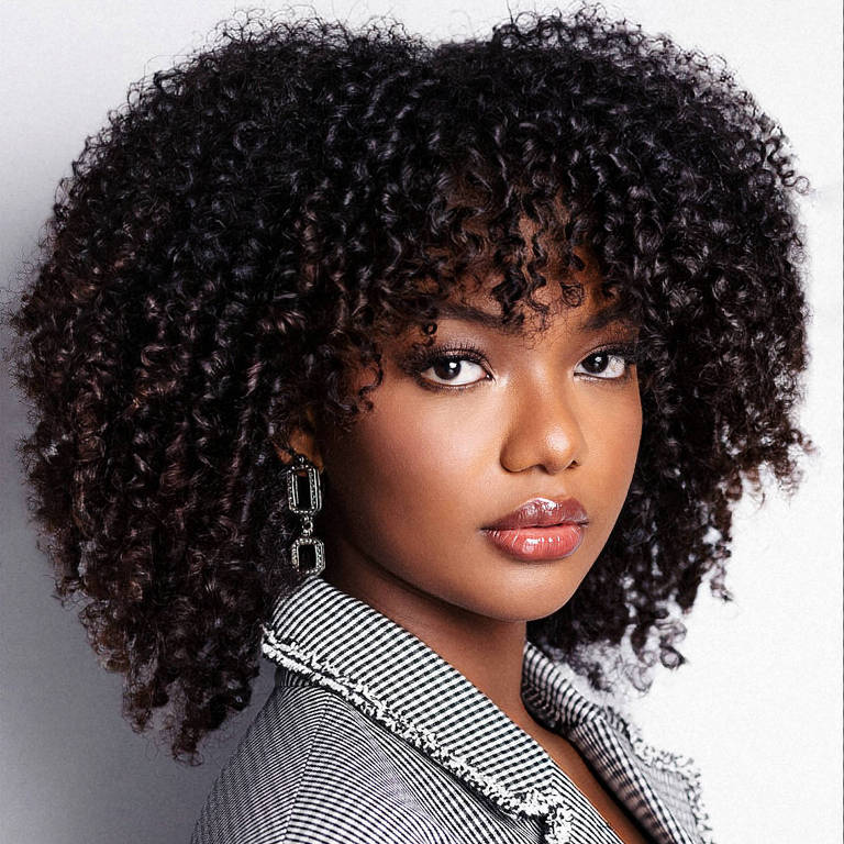 A imagem mostra uma mulher com cabelo cacheado e volumoso, usando um blazer listrado em preto e branco. Ela está olhando diretamente para a câmera, com um leve sorriso nos lábios. A iluminação é suave, destacando suas características faciais e o brilho de sua pele.