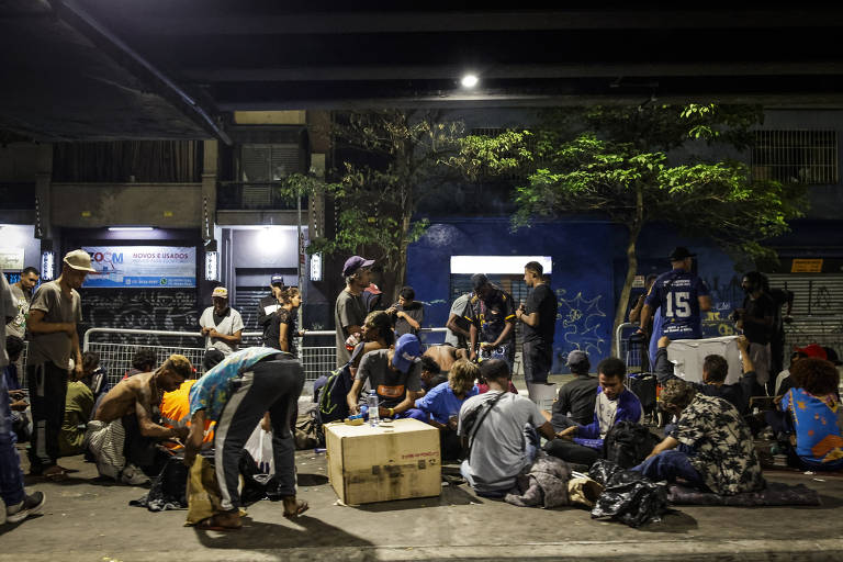 A imagem mostra um grupo de pessoas reunidas em uma área urbana à noite. Algumas pessoas estão sentadas no chão, enquanto outras estão em pé. Há caixas e objetos espalhados pelo chão. O ambiente parece ser uma rua, com iluminação artificial e árvores ao fundo. As pessoas estão vestidas de maneira casual e parecem estar interagindo entre si.