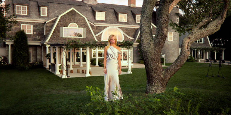 A imagem mostra uma mulher em pé em um gramado verde, vestindo um vestido longo e elegante. Ao fundo, há uma grande casa de estilo arquitetônico tradicional, com telhado de duas águas e janelas amplas. A casa é cercada por árvores e arbustos, criando um ambiente natural e tranquilo.