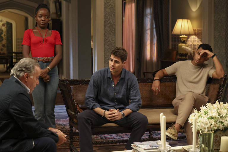 A imagem mostra um grupo de quatro pessoas em um ambiente interno. Um homem mais velho, com cabelo grisalho, está sentado de frente para os outros, que estão em um sofá. Um jovem com cabelo castanho e camisa azul está sentado no centro, parecendo pensativo. Ao lado dele, um homem com camiseta clara está com a mão na cabeça, demonstrando preocupação. Uma mulher com cabelo curto e blusa vermelha está em pé, observando a cena. O ambiente é bem iluminado, com uma mesa de centro decorada com flores.