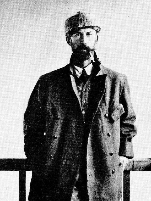 A imagem mostra um homem de pé, vestido com roupas de explorador, incluindo um casaco e um chapéu. Ele tem uma barba e um olhar sério. O fundo é claro, e ele está posicionado em frente a uma estrutura que parece ser uma varanda ou uma grade.