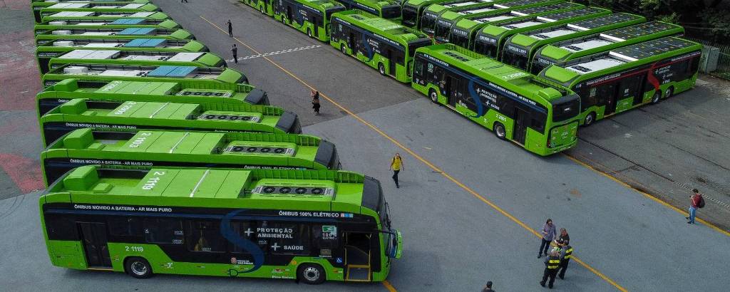 A imagem mostra uma grande quantidade de ônibus estacionados em um pátio. Os ônibus são predominantemente verdes e estão organizados em filas. Algumas pessoas estão caminhando entre os ônibus, e ao fundo, há mais ônibus estacionados. A área é cercada por árvores e vegetação.