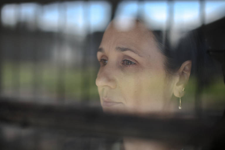 A imagem mostra uma mulher com expressão pensativa, observando através de uma grade. Seu rosto é visível em primeiro plano, com um fundo desfocado que sugere um ambiente externo. A luz natural ilumina seu rosto, destacando seus traços e a profundidade de seu olhar.