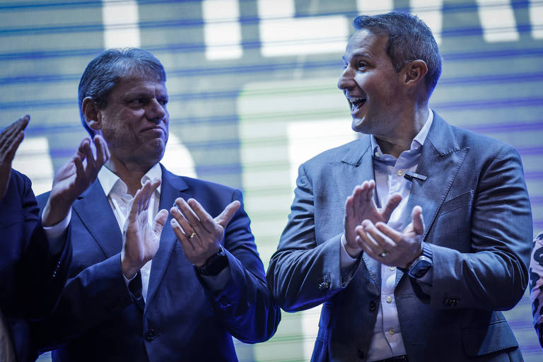 A imagem mostra dois homens em um evento, ambos aplaudindo. O homem à esquerda tem cabelo grisalho e usa um terno escuro, enquanto o homem à direita tem cabelo curto e usa um paletó claro. O fundo é iluminado com luzes que criam um efeito visual. Ambos parecem estar em um momento de celebração.