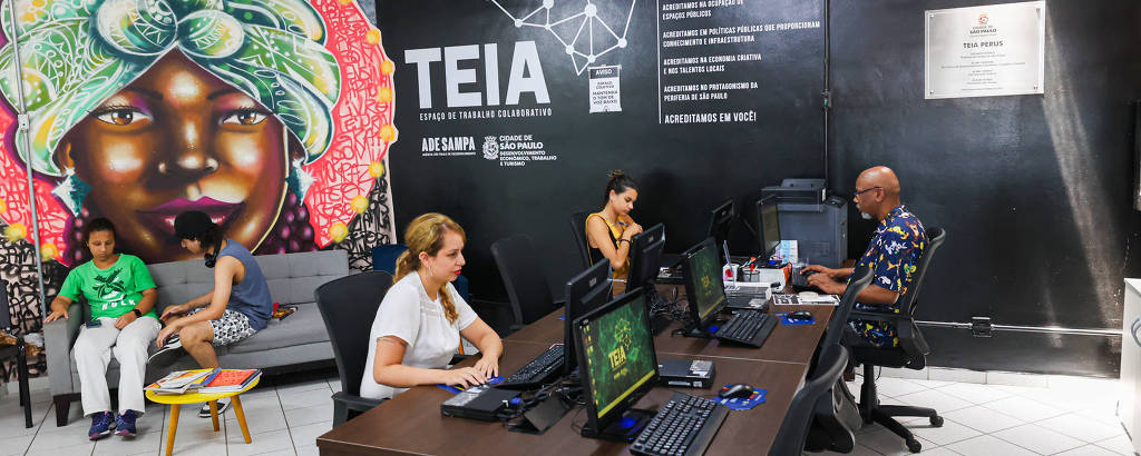 A imagem mostra um ambiente de trabalho colaborativo com várias pessoas. No fundo, há um mural colorido de uma mulher com cabelo verde e adornos. À frente, duas pessoas estão sentadas em mesas de trabalho com computadores, enquanto outras duas estão em um sofá. O ambiente é bem iluminado e possui piso de cerâmica branca.