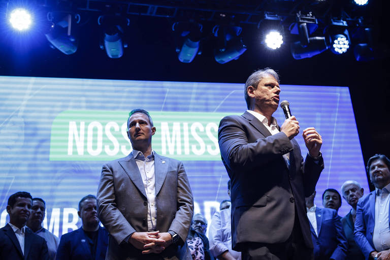A imagem mostra um evento político com várias pessoas em um palco. Um homem em destaque, vestido de terno escuro, está falando ao microfone. Ao seu lado, um homem em terno claro observa. Atrás deles, há um grupo de homens em trajes formais, todos olhando para o orador. O fundo é iluminado com luzes azuis e brancas, e há um telão com a frase "NOSSA MISSÃO" visível.