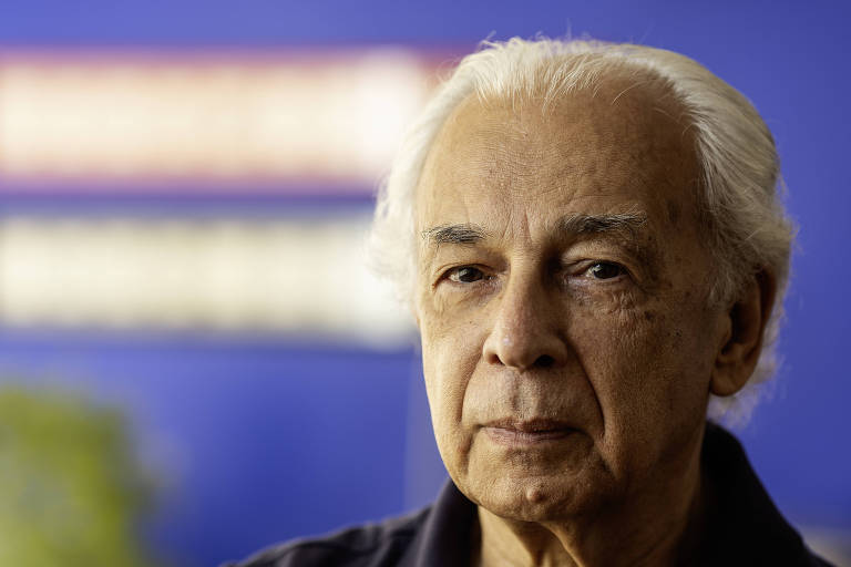 Um homem idoso com cabelo grisalho e pele clara está olhando diretamente para a câmera. Ele tem uma expressão séria e está vestindo uma camisa escura. Ao fundo, há uma parede azul com texto desfocado em cores claras.
