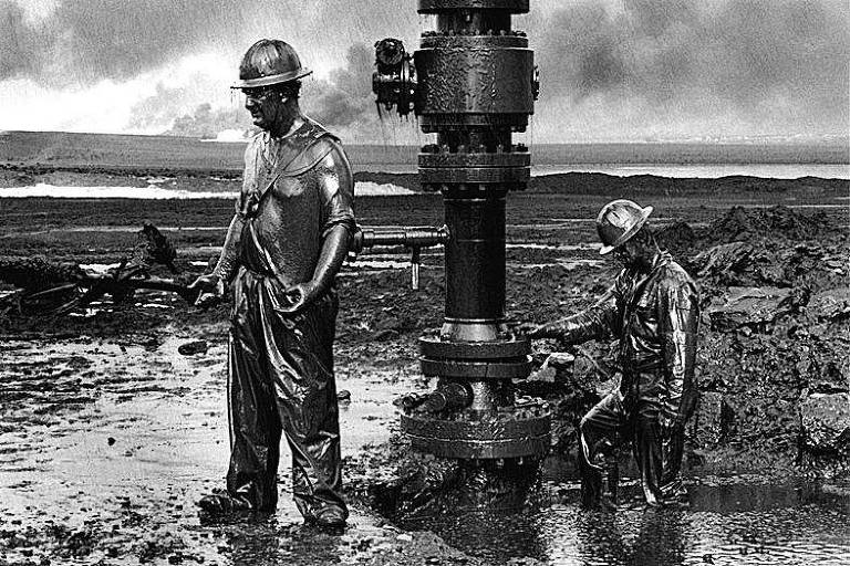 A imagem em preto e branco mostra dois trabalhadores em um poço de petróleo. Ambos estão cobertos de óleo, com roupas sujas e chapéus de segurança. Um dos trabalhadores está em pé, com as mãos nos quadris, enquanto o outro está agachado, manipulando uma válvula no equipamento de perfuração. O fundo apresenta um cenário industrial com fumaça e um horizonte distante.