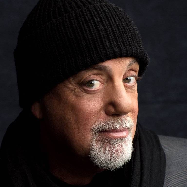 Um homem com cabelo grisalho e barba, usando um gorro preto e um cachecol escuro. Ele está olhando para a câmera com uma expressão amigável, em um fundo escuro.
